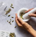 A white mortar and pestle with cardamom pods being crushed in side while a clear plastic bag labeled 'Cardamom' sits beside it.