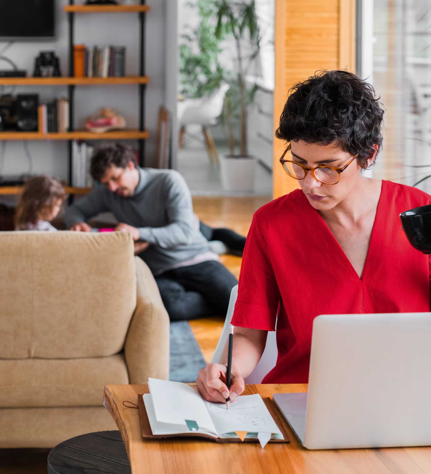Woman working on laptop in living room with family relaxing behind her 