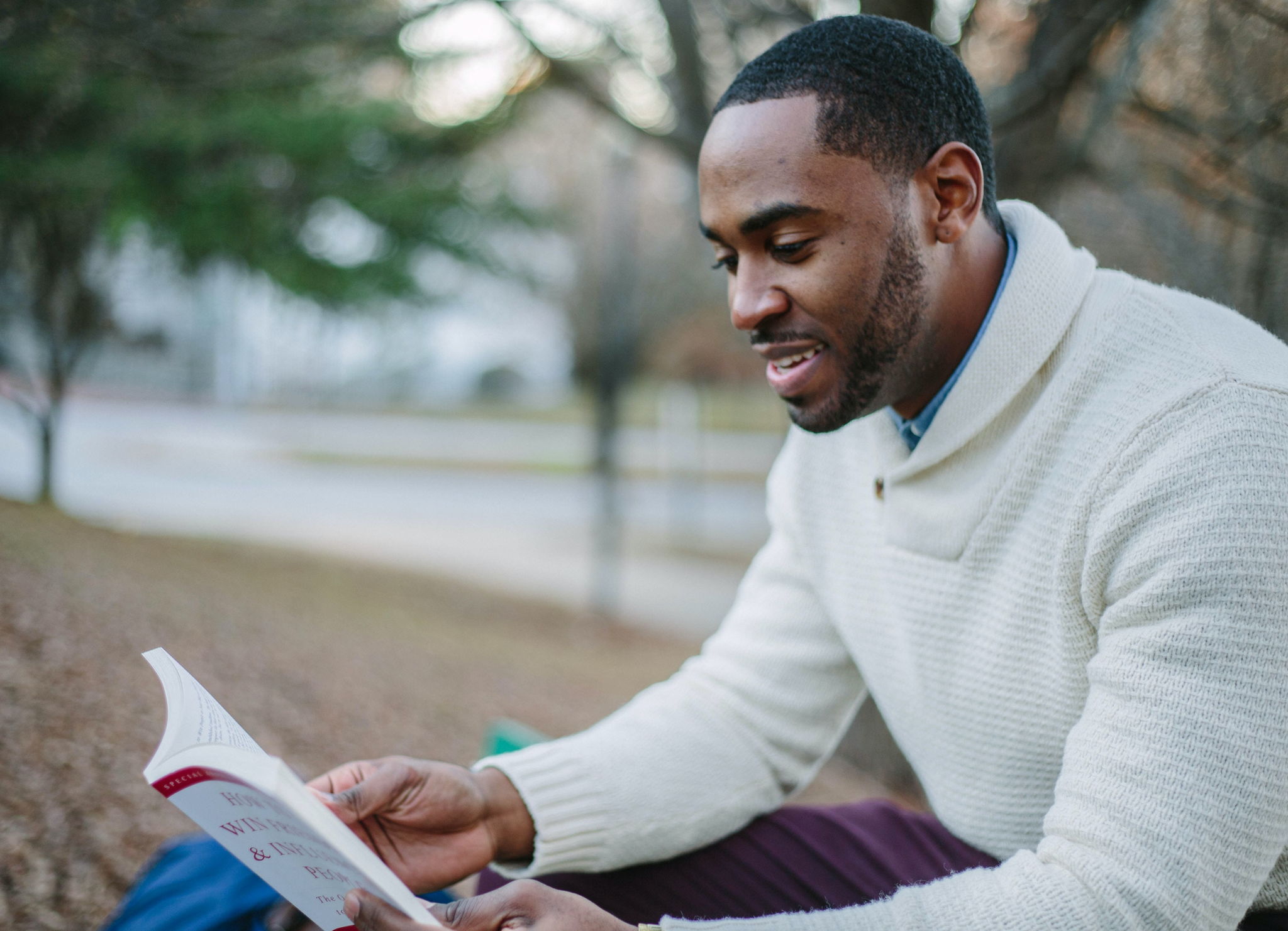 An attractive black man sits in a park and smiles while he reads a book.