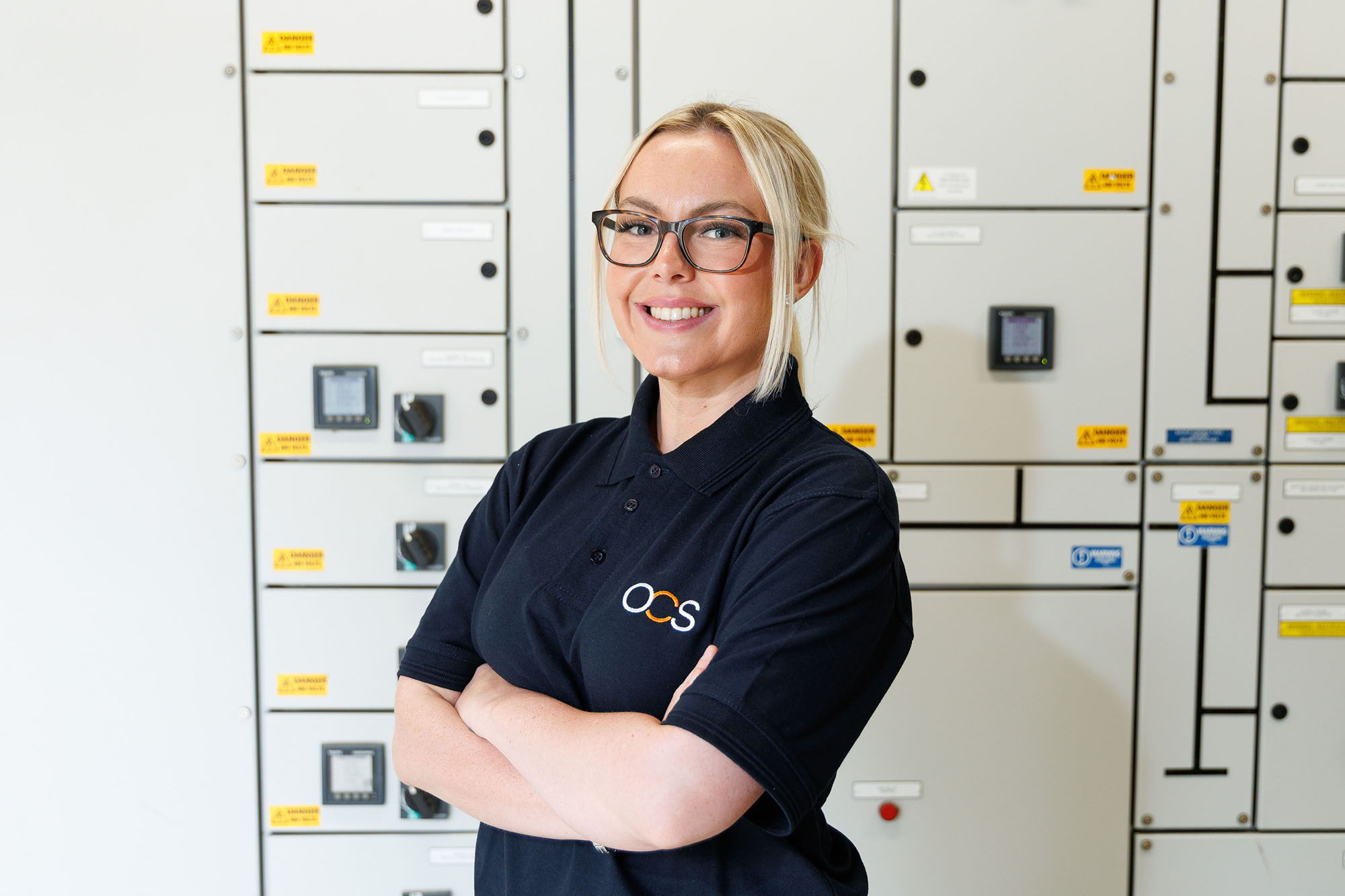 A woman wearing glasses and an OCS-branded navy polo shirt stands smiling with arms crossed in front of a wall of electrical control panels.