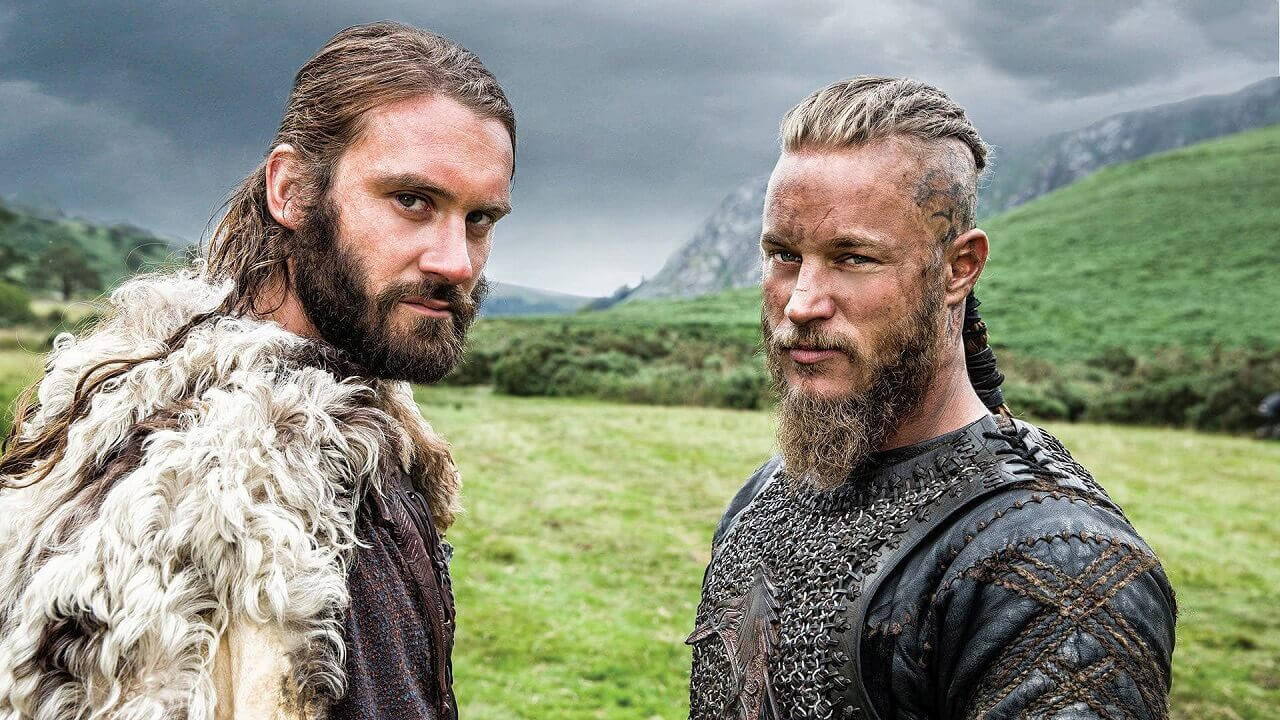 Ragnar and Athelstan with their traditional viking outfit looking intently at the camera with hills behind them.
