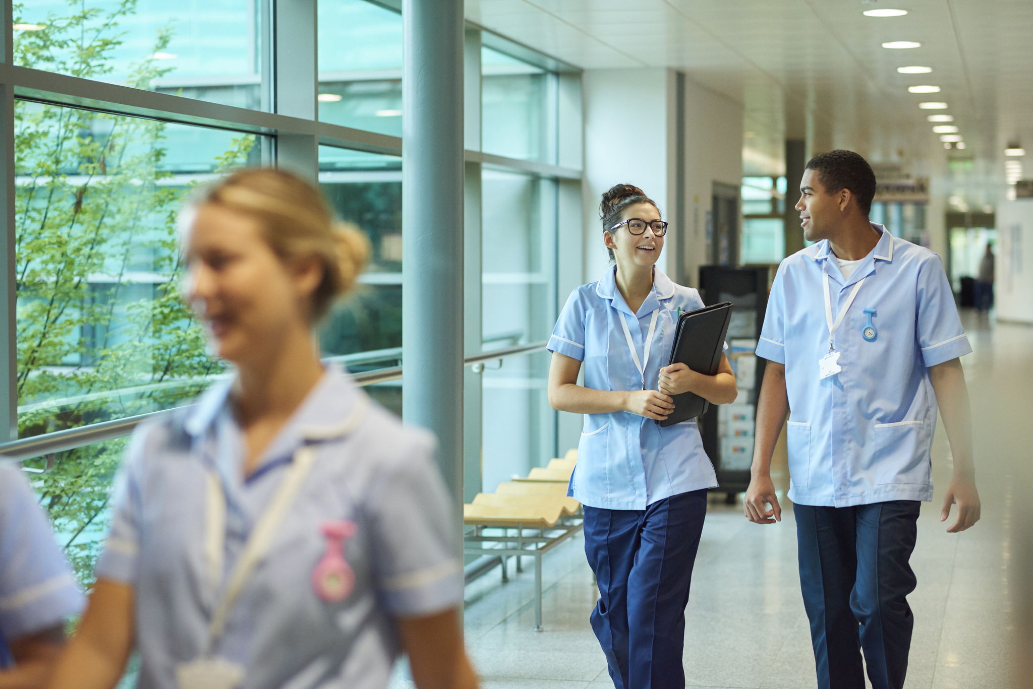 A group of nurses in light blue uniforms walk and talk in a brightly lit hospital corridor with large windows and greenery visible outside.