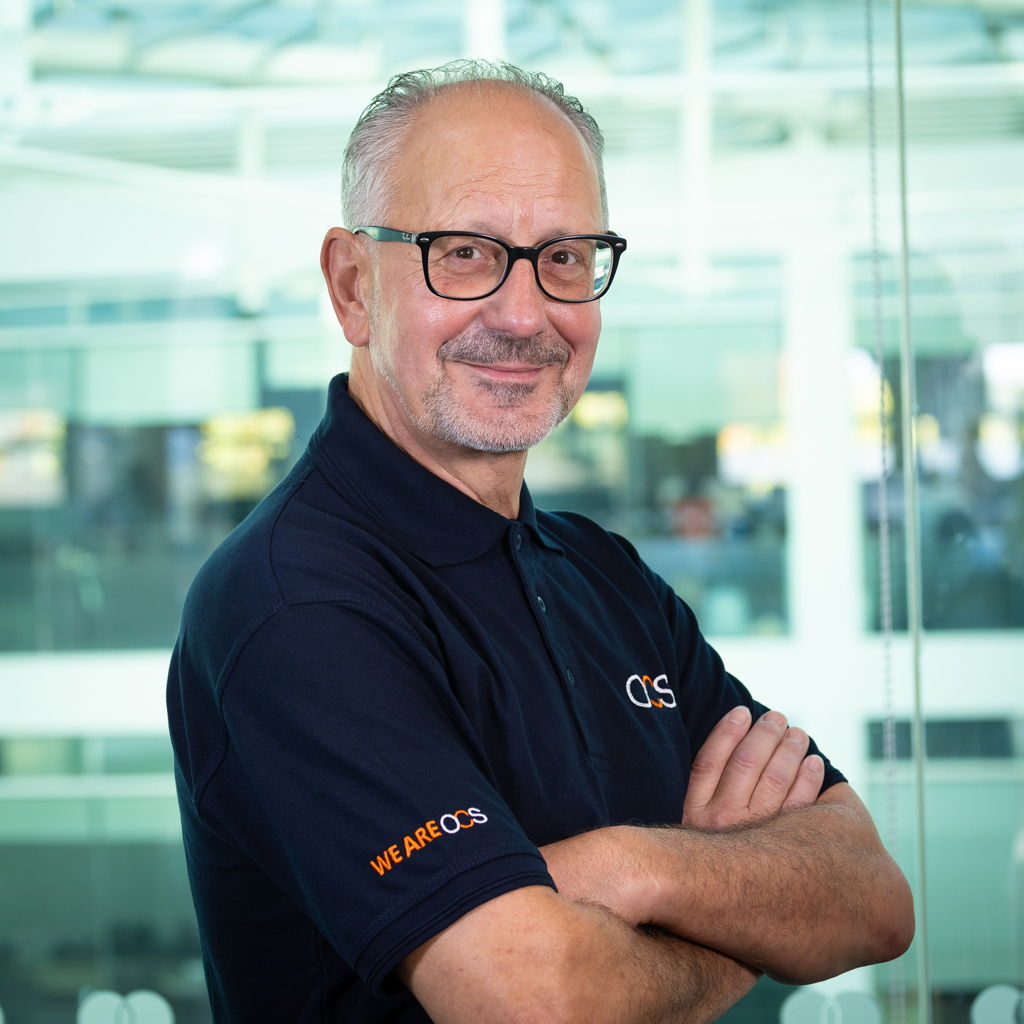 A smiling older man with glasses and a short beard stands with arms crossed, wearing a navy blue polo shirt with OCS and WE ARE OCS logos, in a modern office setting with large windows.