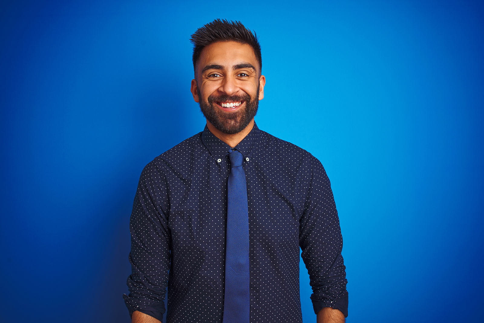 An attractive latin guy wearing a button up and tie smiles for the camera against a simple background.