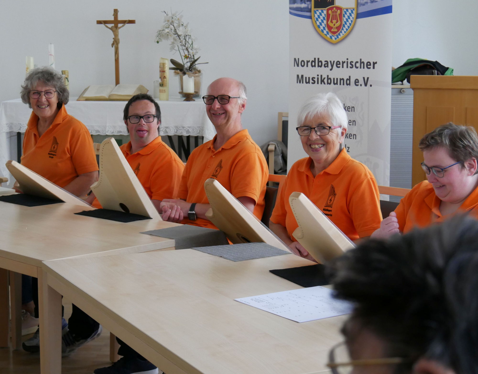 Drei Frauen und zwei Männer mit und ohne Behinderung in einheitlichen orangenen T-Shirts sitzen an einem Tisch nebeneinander und musizieren auf Veeh-Harfen. 