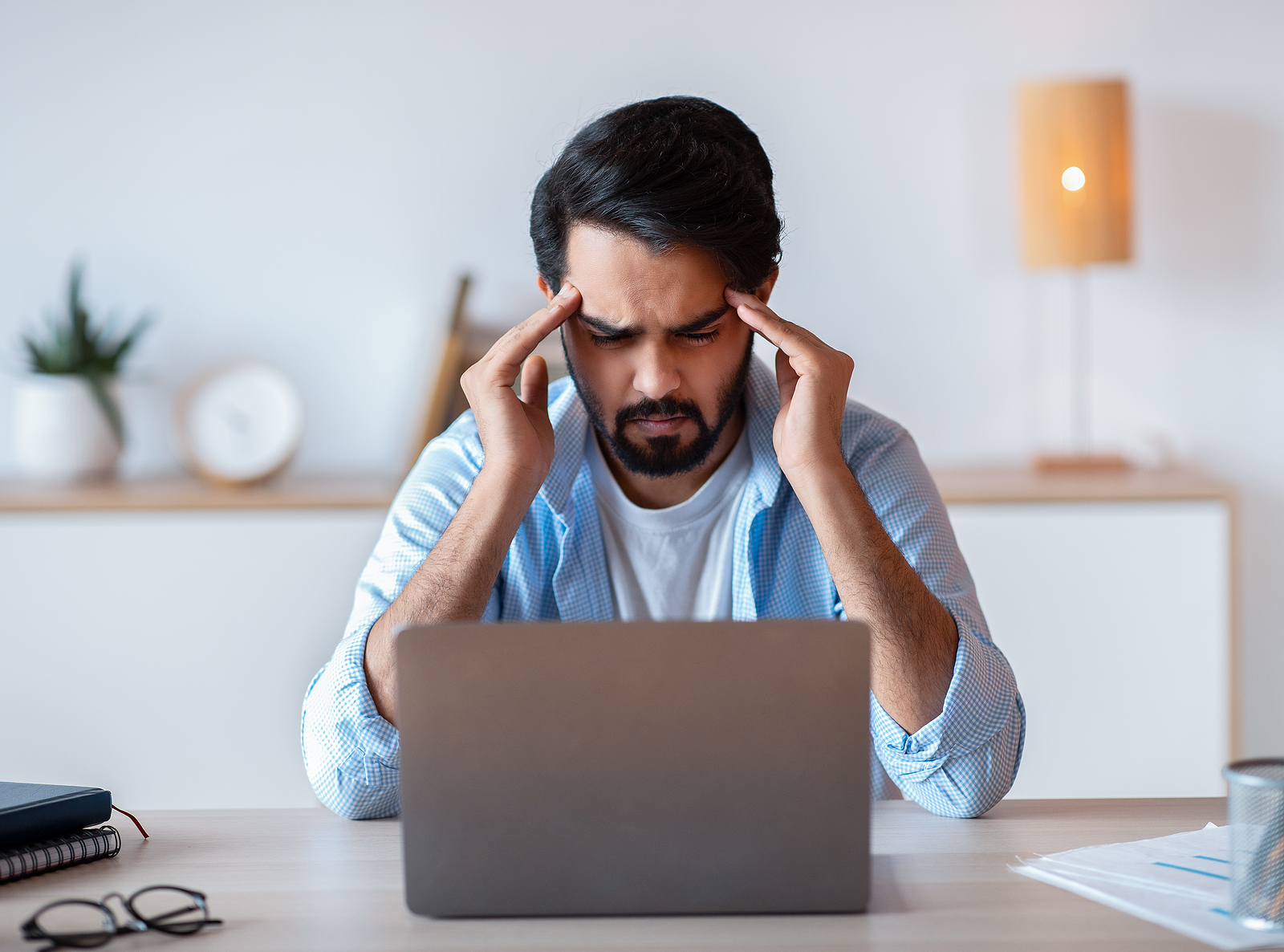 A man sits on his desk looking at his laptop with with frustration with his hands on his temples.