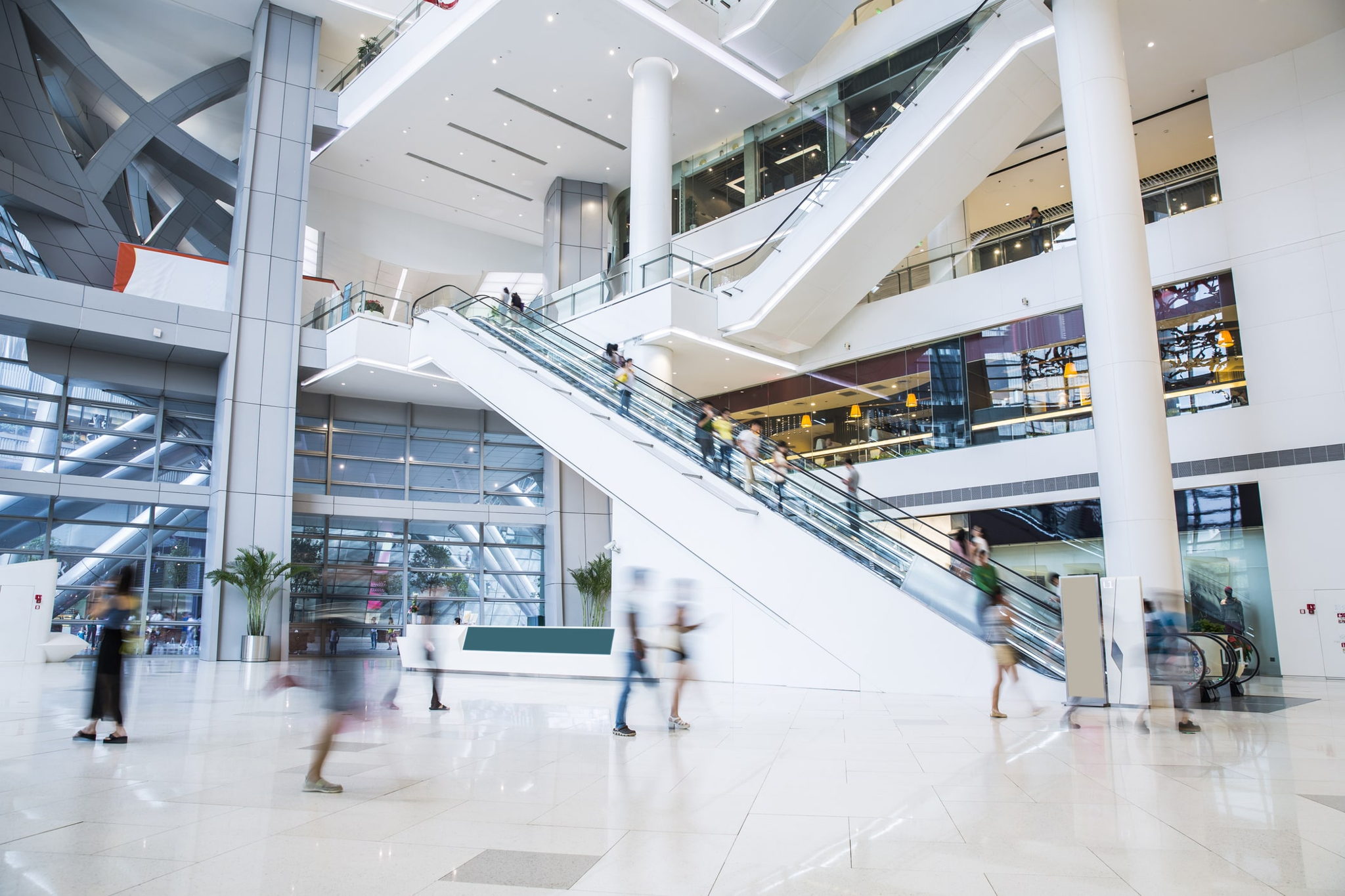 Bright, modern shopping mall interior with people walking and riding escalators. The scene features multiple floors, glass railings, white pillars, and shops in the background, creating a spacious and bustling atmosphere.