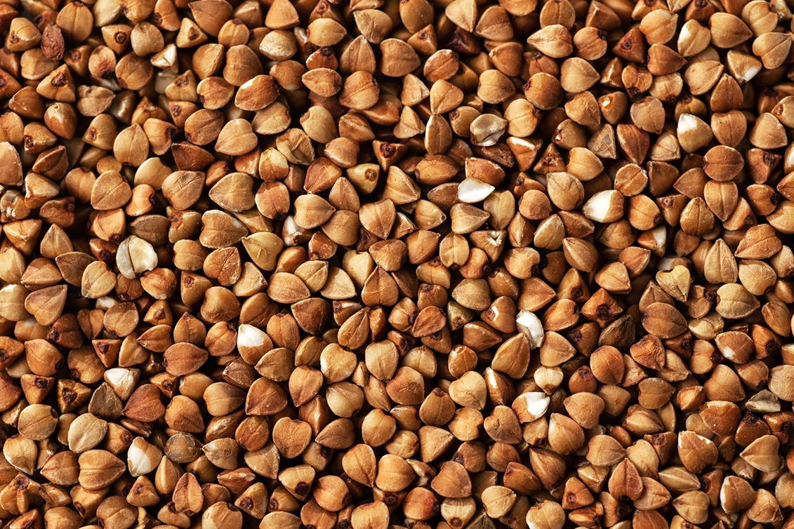 A close-up, top-down photograph filling the frame with raw, light brown buckwheat groats, showcasing their distinct three-sided triangular shape and smooth, matte texture.