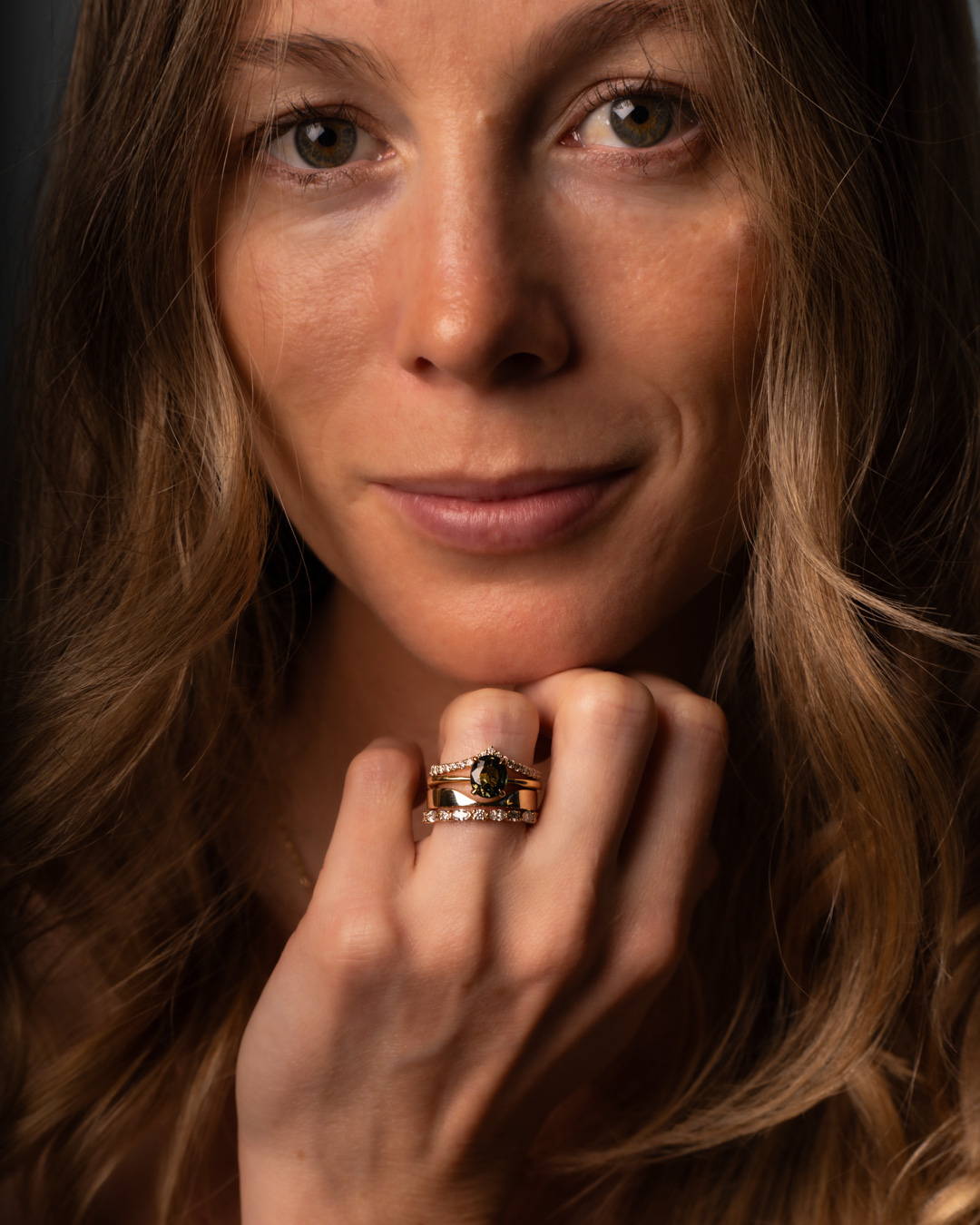 Smiling model wearing a ring stack featuring two diamond bands, a curved gold band, and a solitaire green sapphire ring.