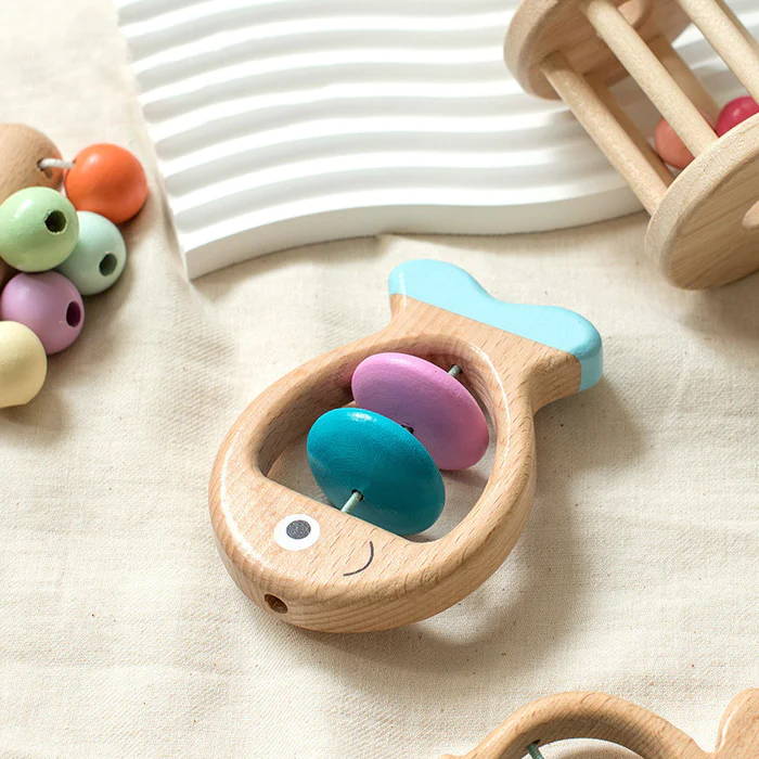 Un kit de hochet Montessori d'InvenToy en forme de poisson, avec des perles colorées bleu sarcelle et violettes, positionnées sur une surface en tissu à côté d'autres hochets en bois.