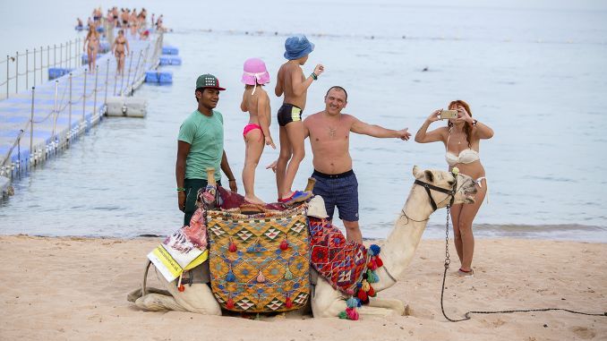 SHARM EL-SHEIKH, EGYPT - MAY 25, 2018  Arab guy along with camel offer their services for tourists on beach near red sea in Sharm El Sheikh, Egypt. Woman is taking pictures of children on a camel — Stock Editorial Photography
