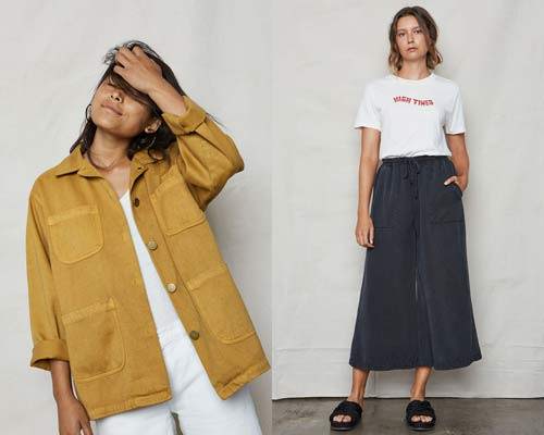 Woman wearing a washed yellow hemp and organic cotton chore workwear jacket with metal buttons and white vest underneath and woman wearing white organic cotton t-shirt with red printed logo reading 'High Times' with dark navy Tencel wide leg pants and black slip on sandals