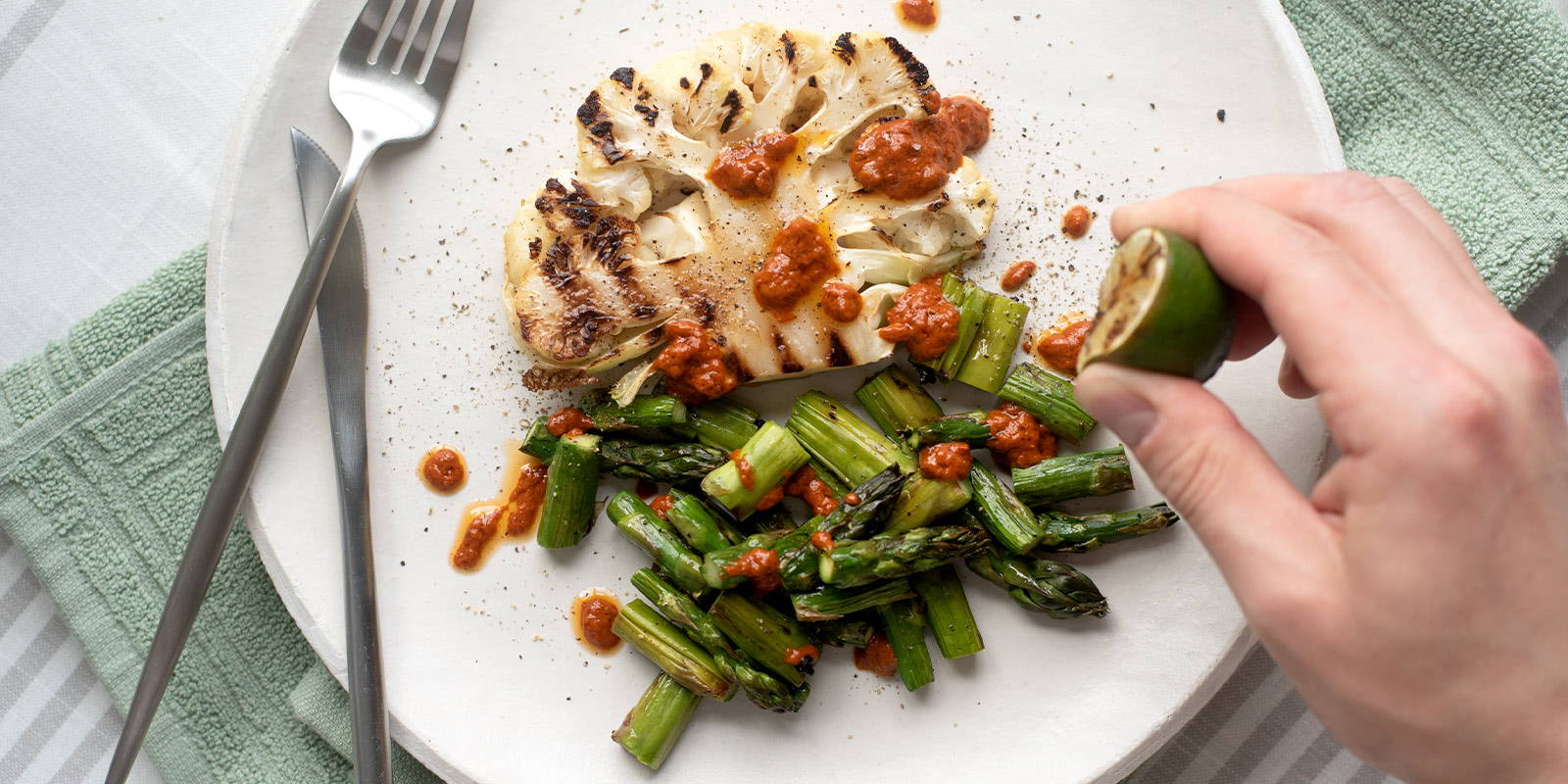 LIme juice is being squeezed over a plate of grilled sliced cauliflower and grilled bite-sizsed asparagus and garnished with harissa.