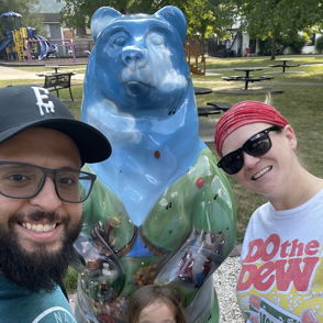 Family smiles around a whimsical bear sculpture in a park, enjoying a joyful and playful outing together.