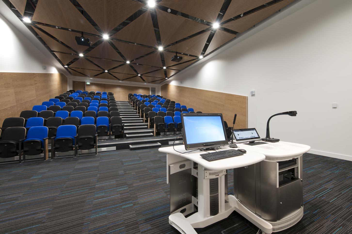 A modern lecture hall with tiered seating, blue and black chairs, and a presenter’s desk in the foreground equipped with a computer, monitor, and document camera. The walls have wood paneling and the ceiling has geometric patterns.