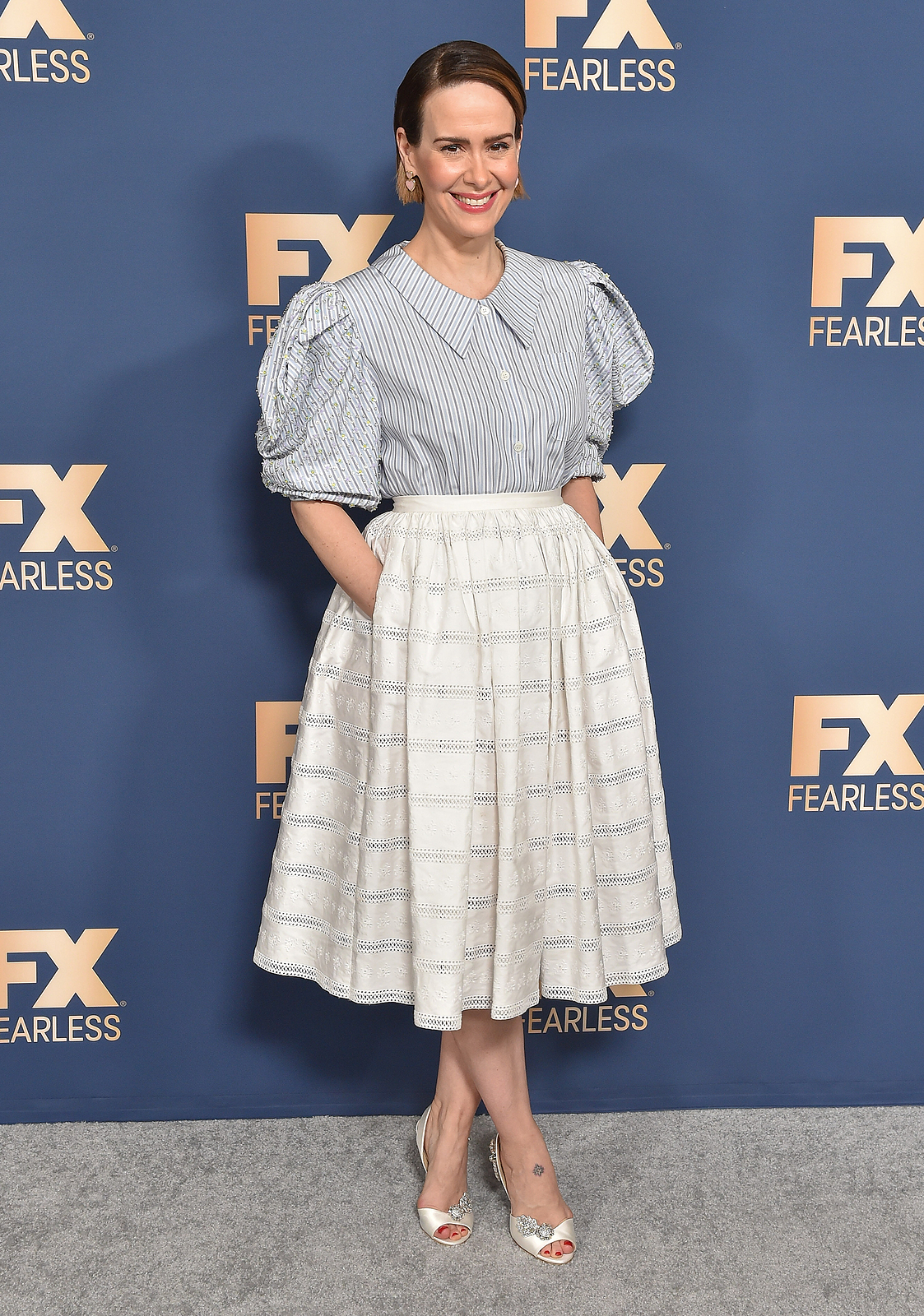 Sarah Paulson at a carpet event with her hands inside her dress pockets and smiling.