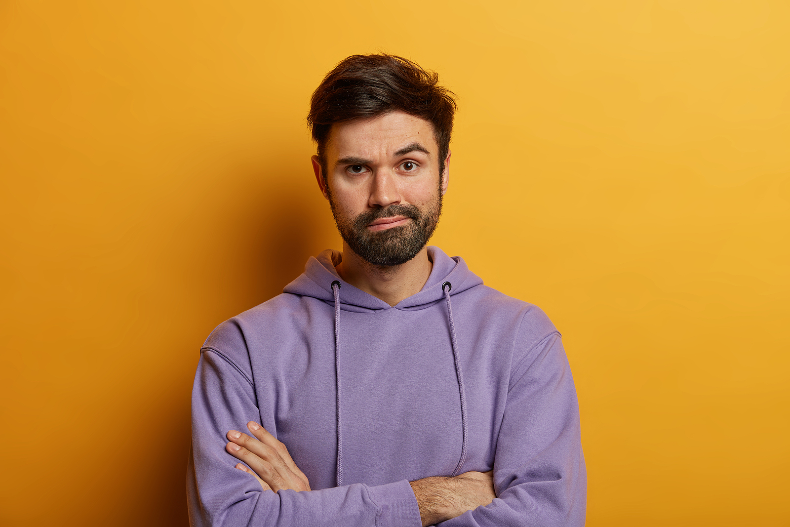 Image of an attractive caucasian man wearing a purple shirt against a plain yellow background, with his arms crossed and an annoyed expression on his face.