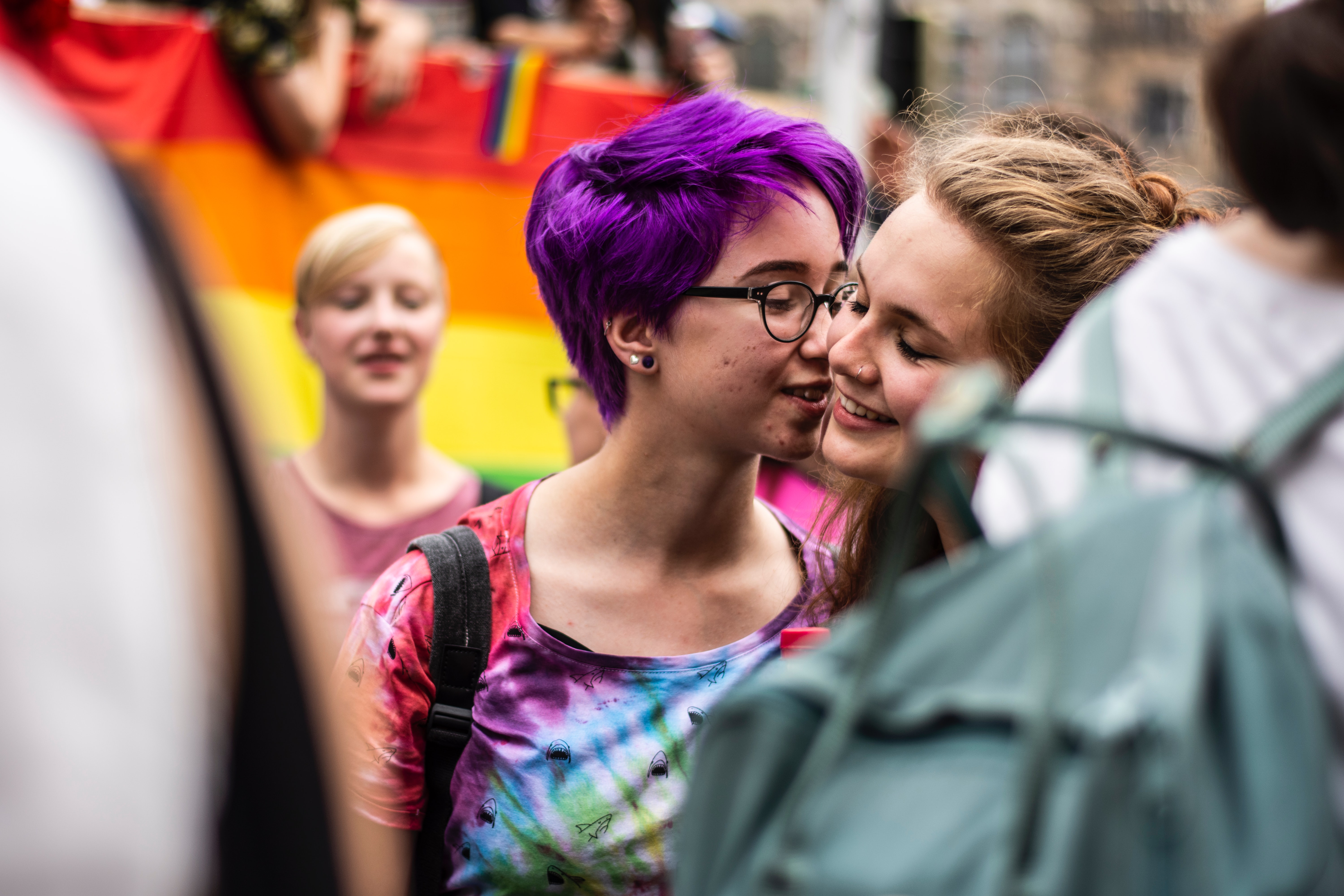 Two women, one with dyed purple hair, share an intimate moment talking during a pride parade.