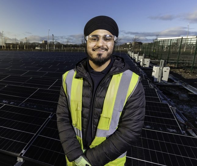 A smiling person wearing a turban, safety glasses, and a high-visibility vest stands in front of rows of solar panels on a clear day.