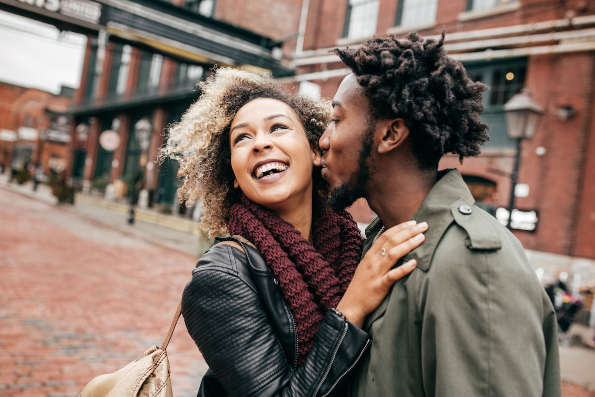 Image of two african american attractive people close together in the street wearing warm clothing. The man is kissing the girls cheek while she smiles coyly looking away.