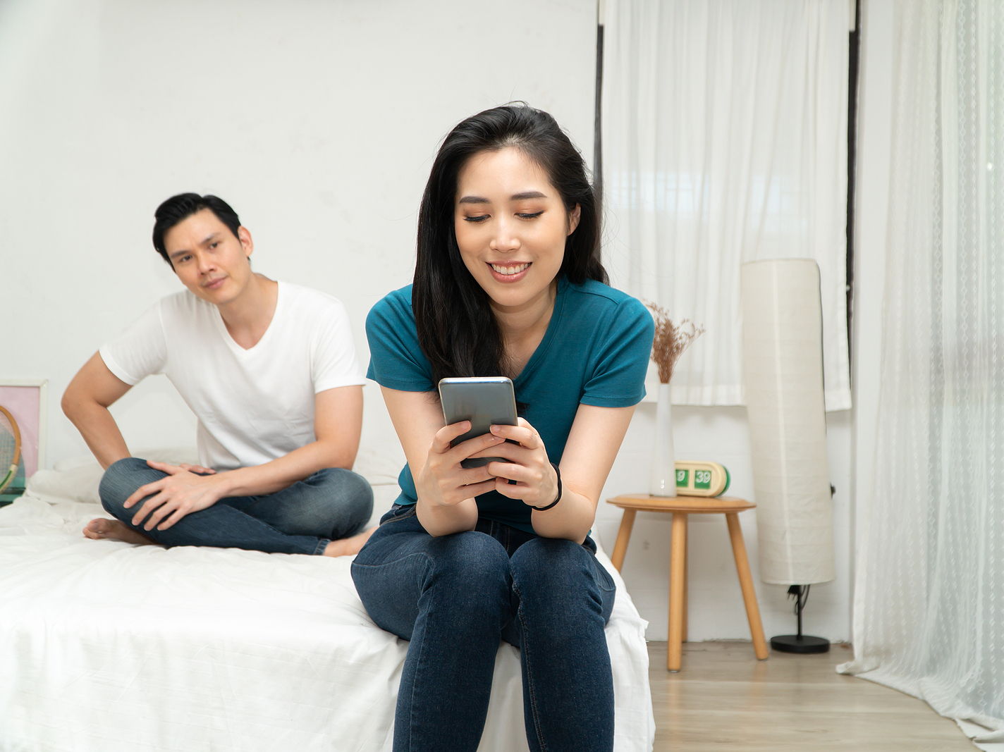 An asian couple sits in bed. The man looks suspiciously at his girlfriend as she looks on her phone smiling.