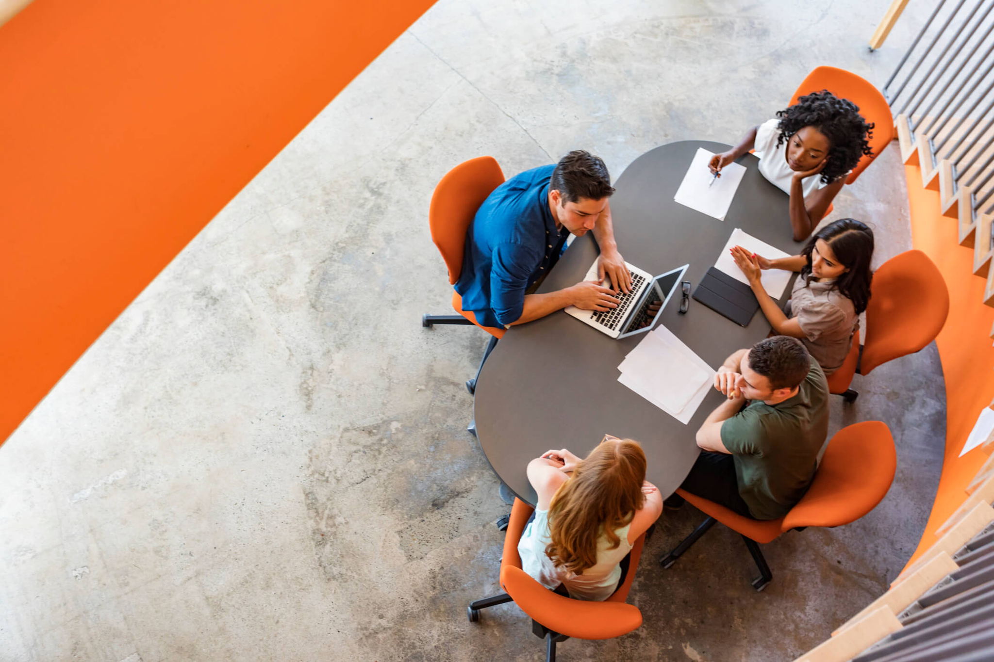 A group of five people are sitting around a table with an open laptop and papers. They appear to be collaborating or discussing work. The setting is modern, with orange chairs and a concrete floor. The photo is taken from above.