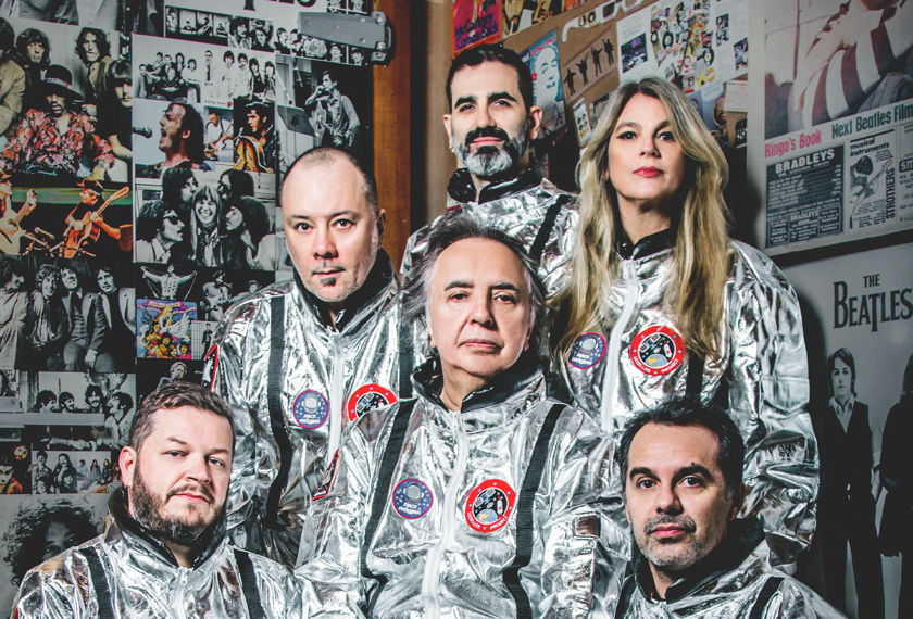 Members of Os Mutantes wearing aluminum spacesuits, behind them are posters of rock bands. 