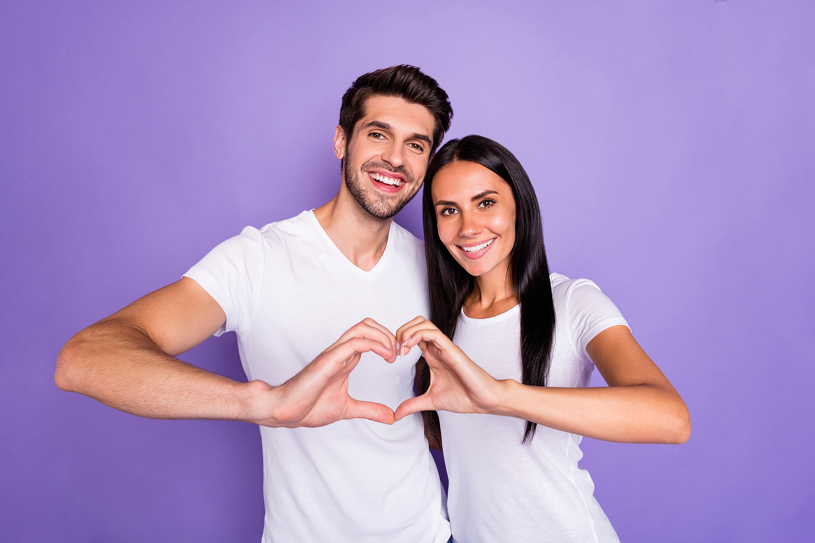Portrait of a man and woman wearing plain t shirts and making a heart shape with one hand each, smiling.