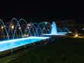 Beleuchteter Garten bei Nacht mit Wasserfontänen und einem langen, rechteckigen Wasserbecken inmitten einer Rasenfläche.