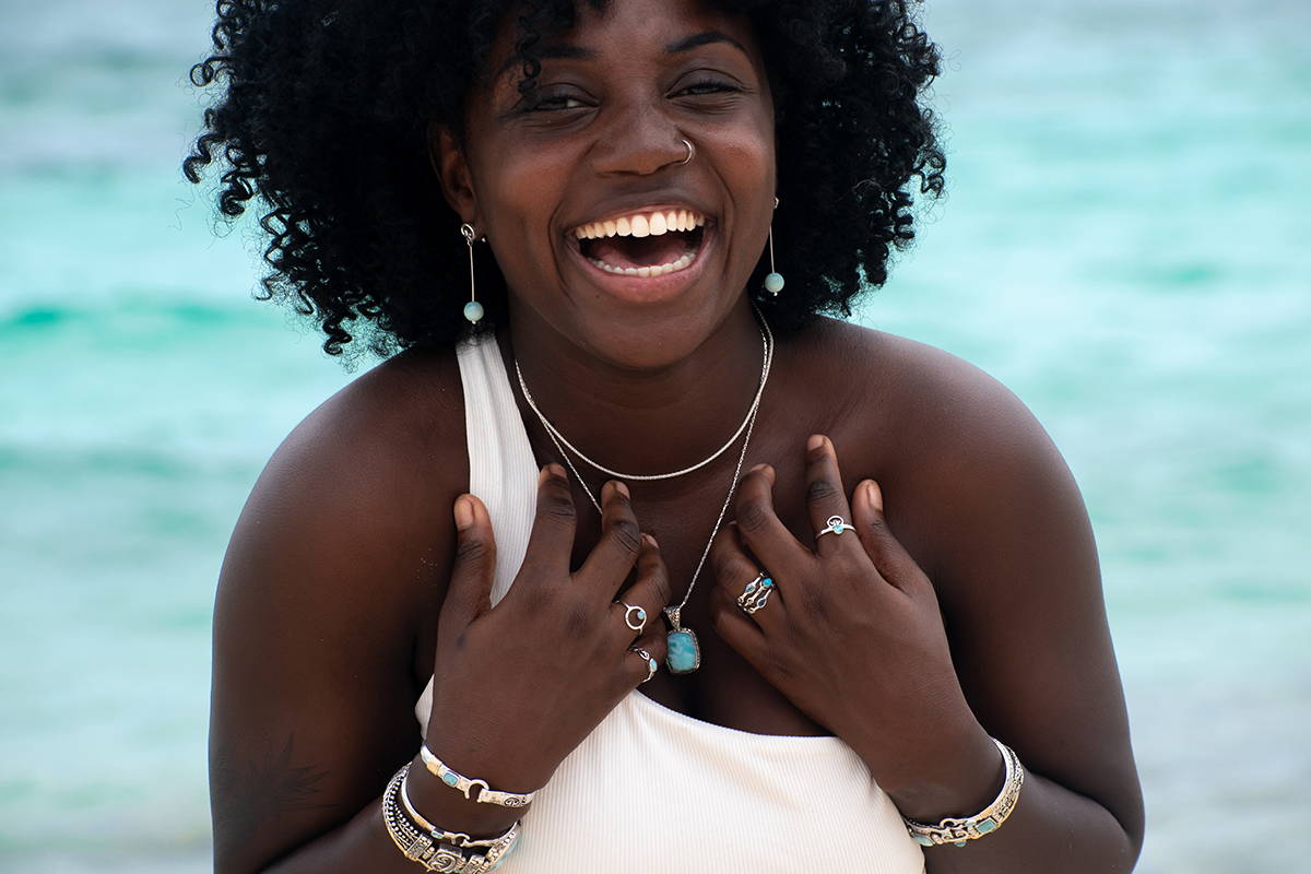 A woman laughing next to the ocean wearing pendants and chains.