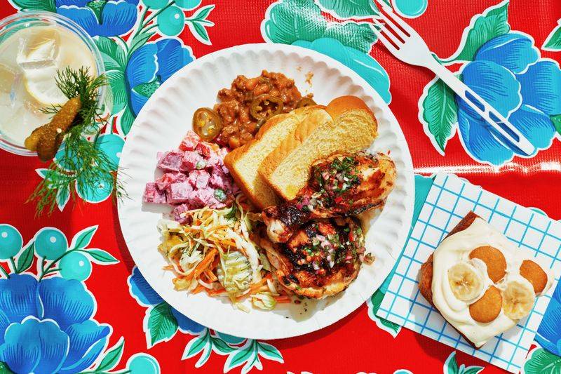 Pink potato salad, pickle coleslaw, burger beans, better-than-beer-can chicken, banana pudding cake, and slices of bread on a plate with a glass of pickle lemonade.