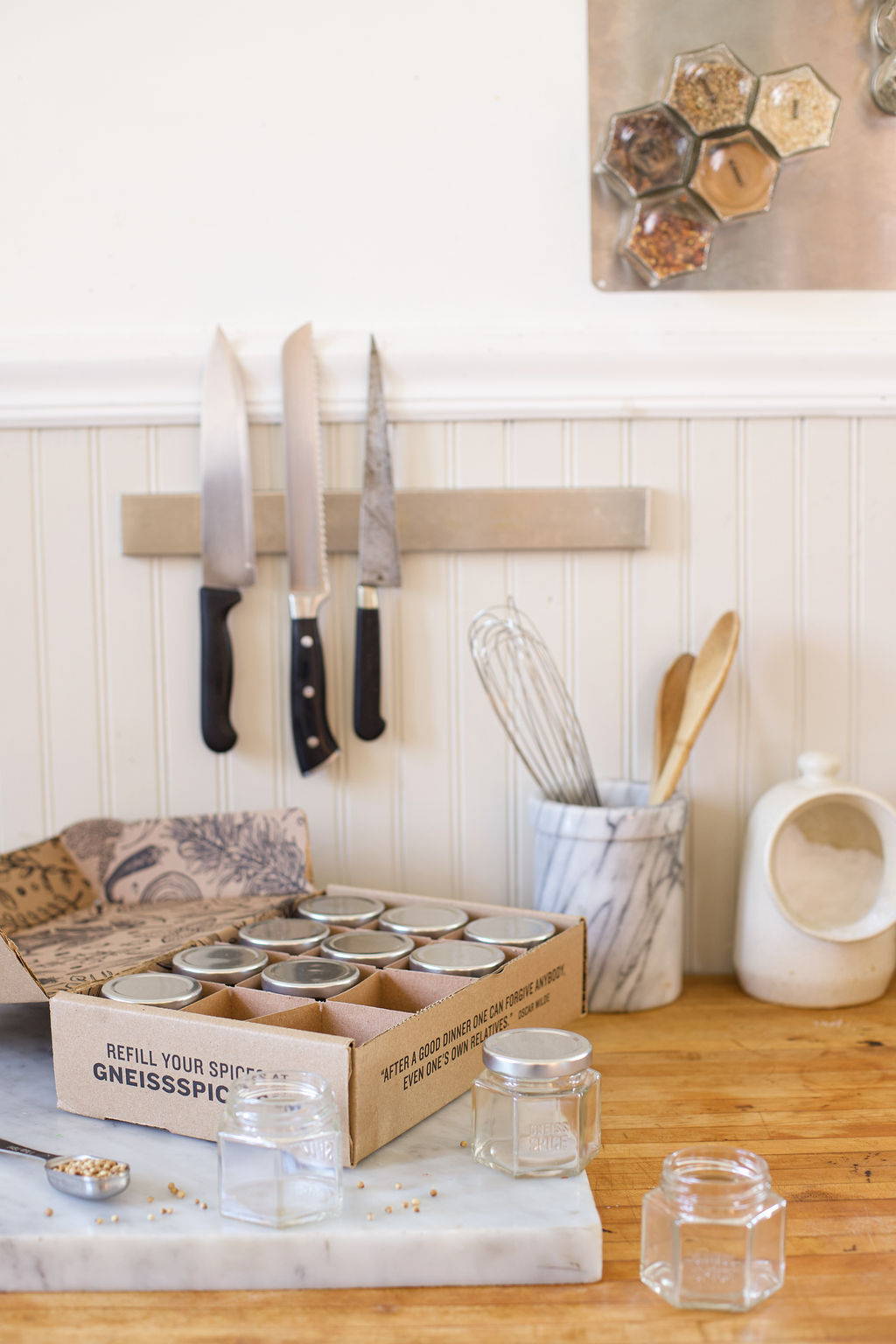 A brightly lit kitchen shows a cardboard box labeled 'Gneiss Spice' filled with glass jars sitting on a marble cutting board.