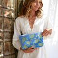 A woman in a white dress standing in speckled sunlight against an old wooden door. She is holding an Elizabeth Scarlett velvet embroidered wash bag in a turtle design, with a mid blue background.