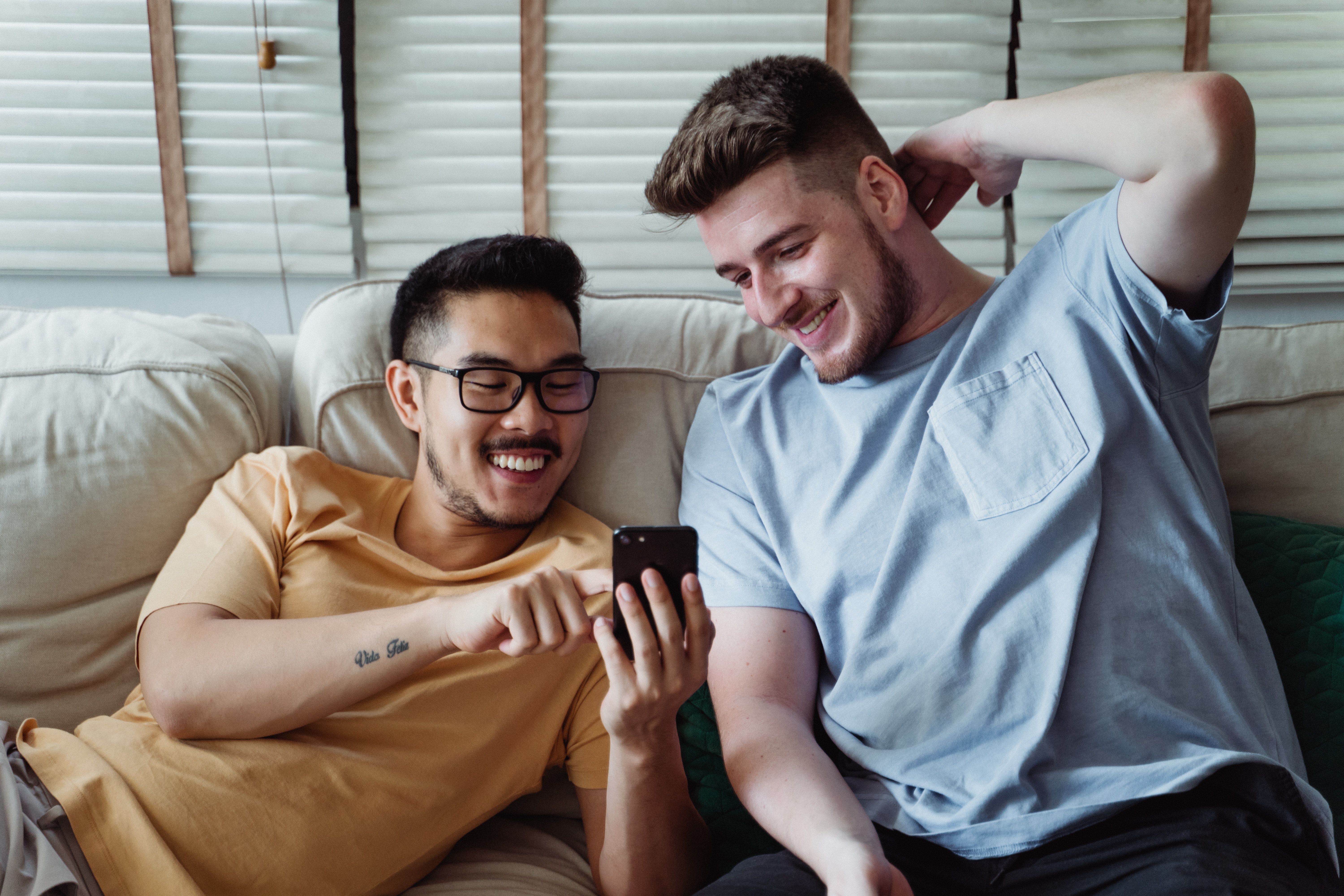 Two guys laugh while looking at a phone together sitting on a couch close to eachother.