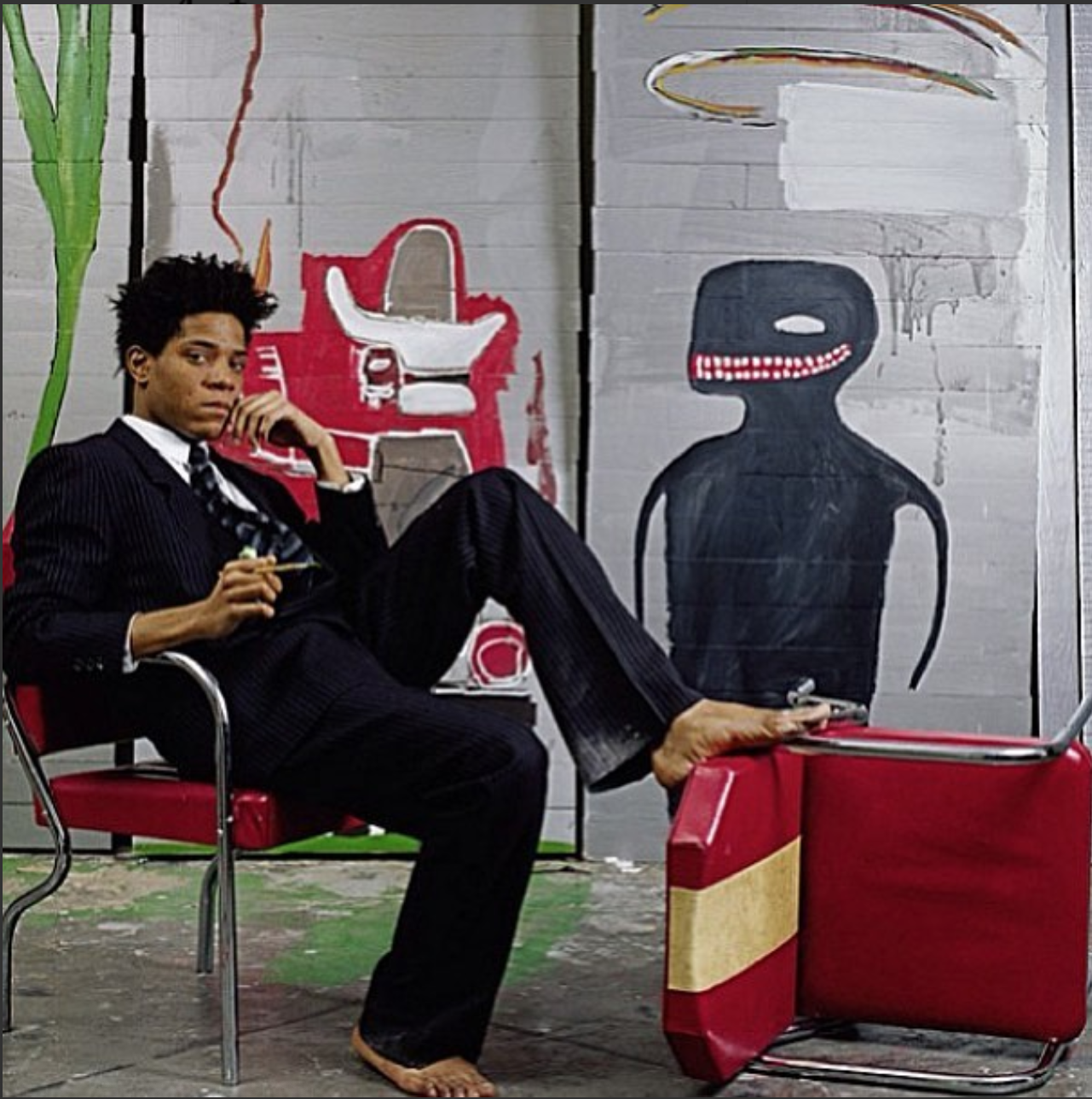 Basquiat sitting on a chair without shoes, with a leg on another chair. Behind his is his art on a wall.
