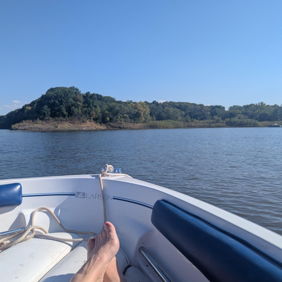 Tranquil boat scene with feet at the edge, calm water, blue sky, and greenery; evokes serenity, freedom, and nostalgia.