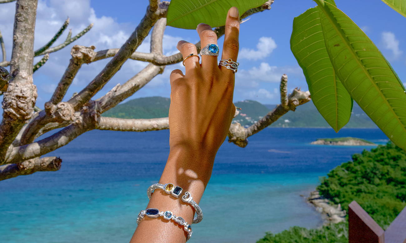 Hand wearing blue sparkling gemstone rings and bracelets while holding a branch with an ocean in the background.
