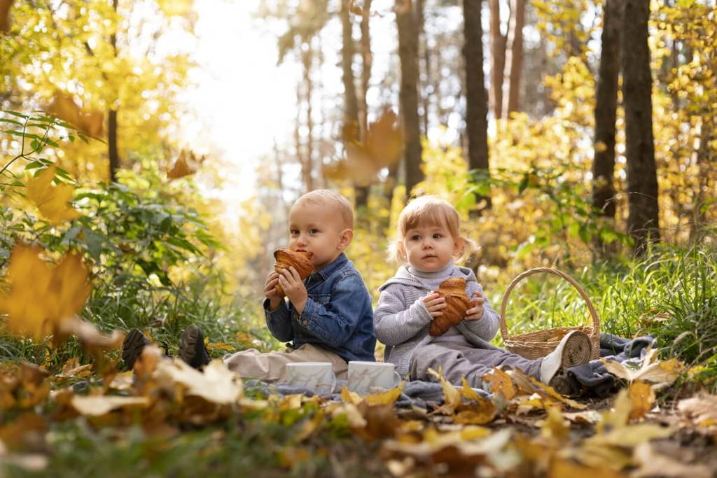 Toddlers sitting in the forest and eating croissants.