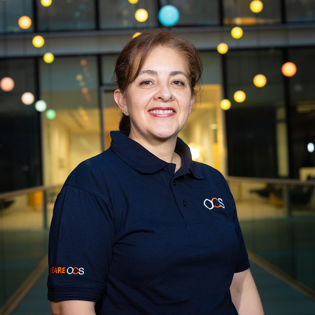 A woman with light brown hair tied back smiles at the camera. She wears a navy blue OCS polo shirt and stands indoors with colorful lights hanging in the background.