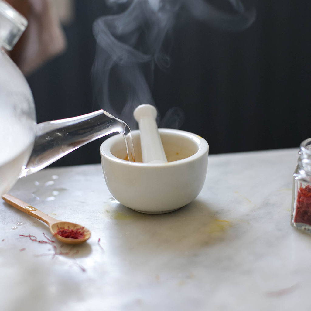 Hot water is poured from a clear glass tea pot into a white mortar and pestle with a small wooden spoon of saffron laying next to it