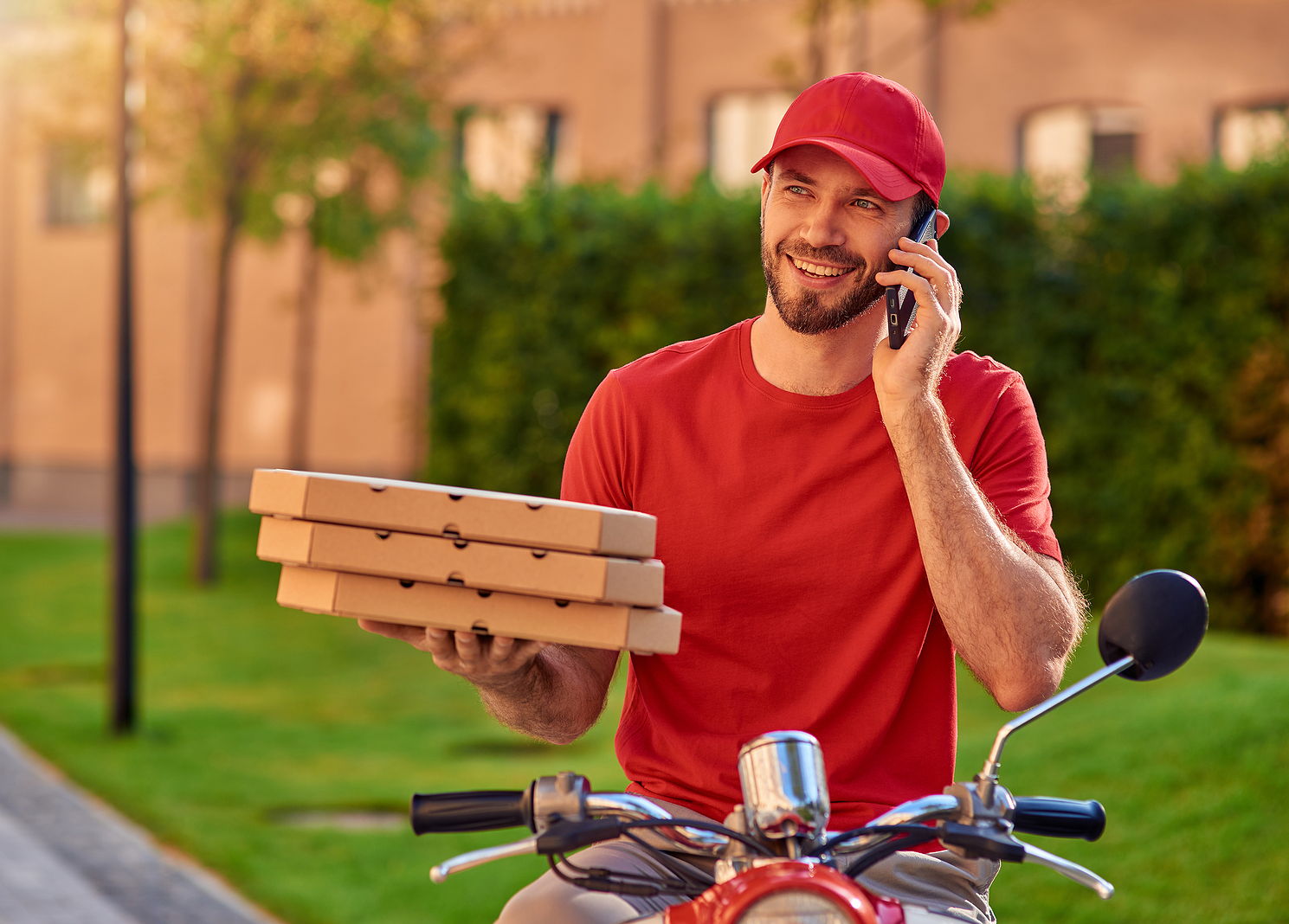 A delivery worker in uniform making a phone call on his bike with 3 pizza boxes in hand.
