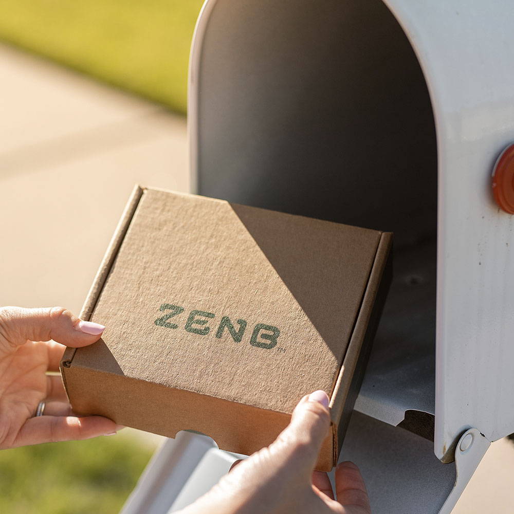 ZENB recycled shipper box is being pulled out of a white mailbox by a woman's hands on a sunny day