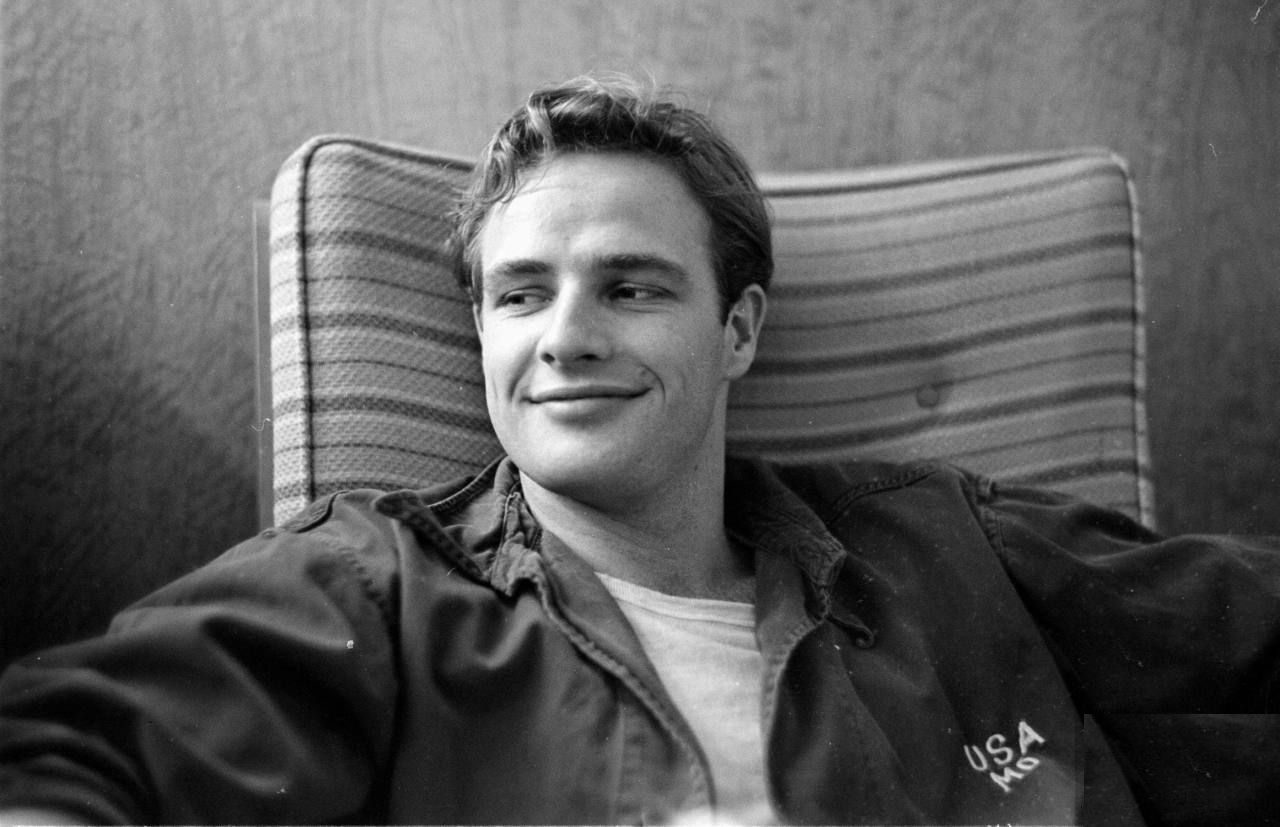 Black and white image of Marlon reclining on a chair and smirking looking at his side.