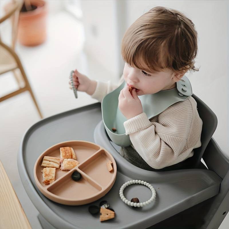 Un tout-petit assis dans une chaise haute se nourrit lui-même de collations provenant d'un coffret cadeau Montessori First Bites divisé par InvenToy. L'enfant porte un bavoir et il y a des jouets à proximité, dans une pièce lumineuse et doucement éclairée.