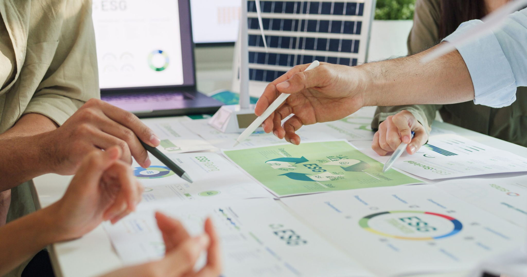 Four people discuss ESG (Environmental, Social, Governance) reports at a table covered with charts and documents. A laptop with graphs and a small solar panel are also visible, highlighting sustainability themes.