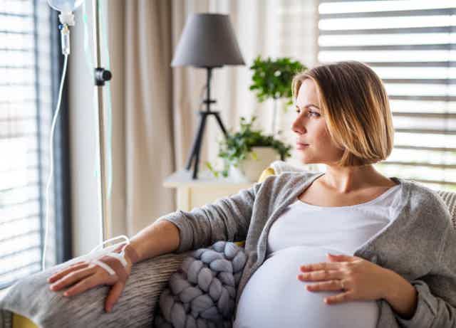 Pregnant woman seated on a couch, gently holding her belly near a window with soft daylight and plants.