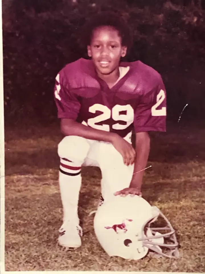 Red-tone color photo of Chris Robinson as a child in Football gear, kneeling on the grass with his hand on his helmet on the ground
