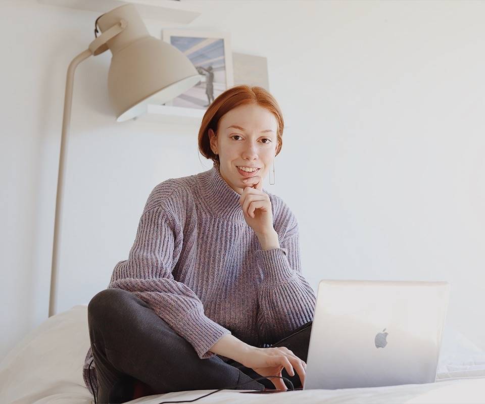 Junge rothaarige Frau sitzt mit Laptop vor sich auf dem Bett und schaut in die Kamera