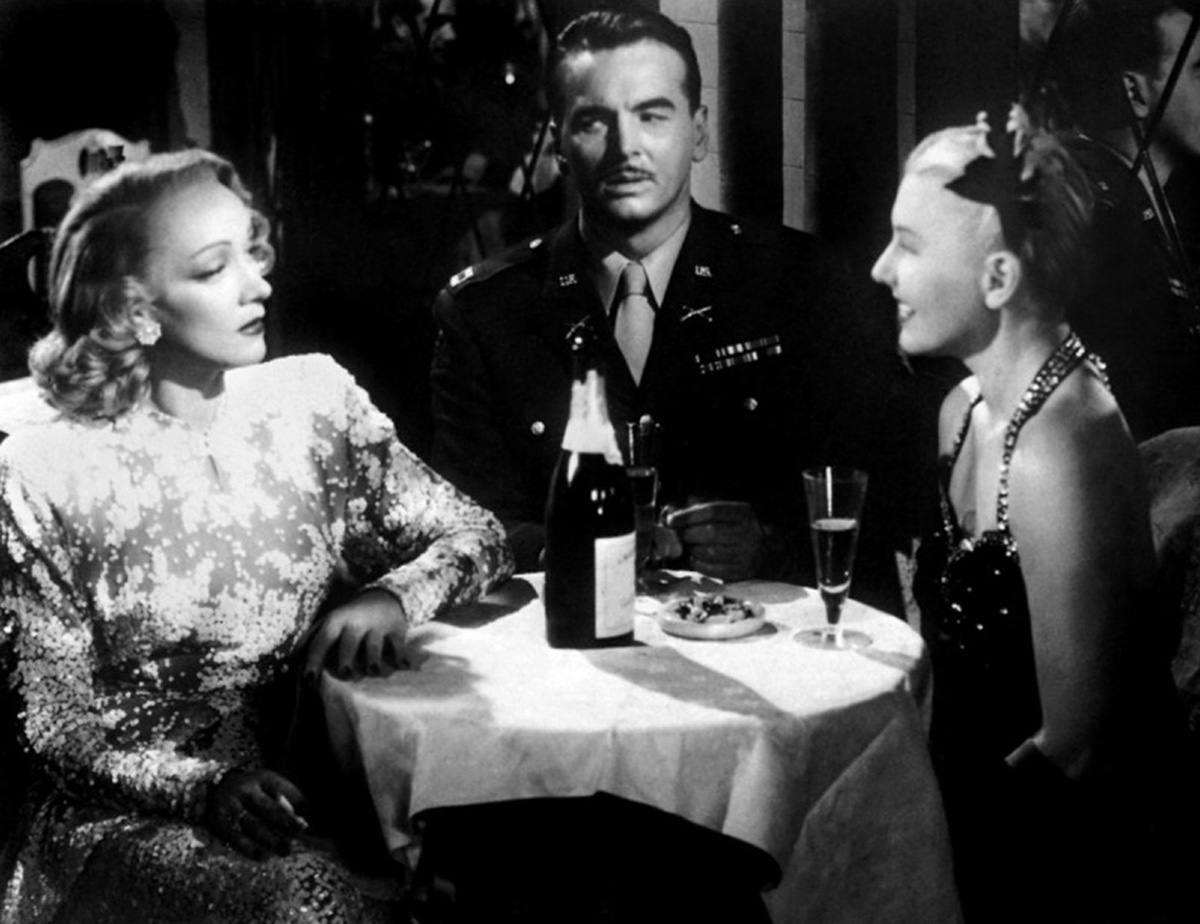 Marlene talking to a woman who is smiling in front of her while a soldier sits next to them at their table.