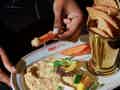 Eine Hand taucht ein Karottenstück in Hummus, serviert mit Gemüse und Brot auf einem dunklen Marmortisch.